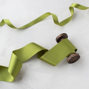 May include: A spool of green satin ribbon with a wooden spool. The ribbon is unraveled and laying on a white textured surface.