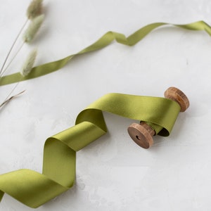 May include: A spool of olive green satin ribbon with a wooden spool. The ribbon is lying on a white textured surface.