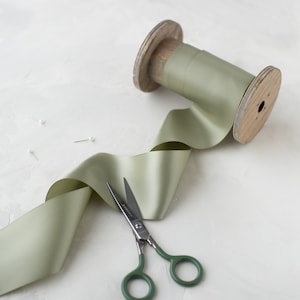 May include: A spool of sage green satin ribbon with a wooden spool and a pair of green handled scissors.