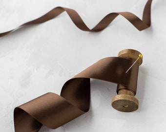 May include: A spool of brown satin ribbon with a gold pin, on a white background.