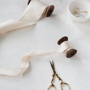 May include: Two spools of white silk ribbon with wooden spools, a small pair of gold scissors, and a small bowl of gold pins on a white surface.
