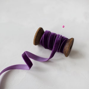 May include: A spool of purple velvet ribbon with a pin holding the ribbon in place. The ribbon is wound around a wooden spool.