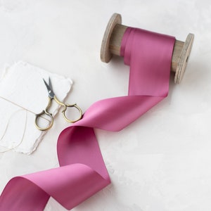 May include: A spool of dusty rose satin ribbon with a wooden spool and a pair of antique silver scissors on a white textured background.
