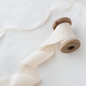 May include: A spool of off-white ribbon with a wooden spool. The ribbon is unraveled and draped over a white surface.