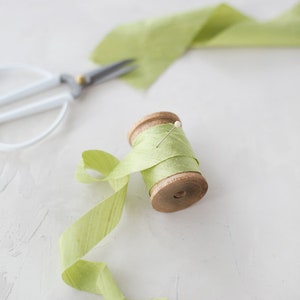 May include: A wooden spool of light green ribbon with a pin in it. The ribbon is unraveled and laying on a white surface.