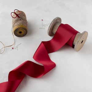 May include: A spool of red satin ribbon on a wooden spool with a length of ribbon extending from the spool. The ribbon is on a white surface with a spool of twine and a pair of scissors.