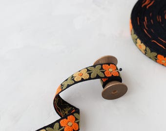 May include: A black fabric ribbon featuring an embroidered floral pattern in shades of orange, yellow, and olive green. The ribbon is wrapped around a small wooden spool, with a portion of the trim extending outward and secured by a pin.