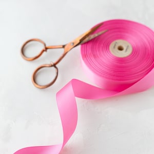 May include: A pink satin ribbon with a spool of ribbon and a pair of gold scissors on a white background.
