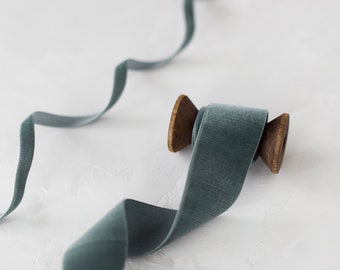 Puede incluir: Una bobina de cinta de terciopelo azul turquesa con una bobina de madera. La cinta está sobre una superficie blanca.