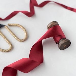 Puede incluir: Un carrete de cinta rojo oscuro con núcleo de madera, parcialmente desenrollado, sobre una superficie blanca. Tijeras doradas en el fondo. La cinta es lisa, probablemente de satén. Ideal para manualidades y envolver regalos.