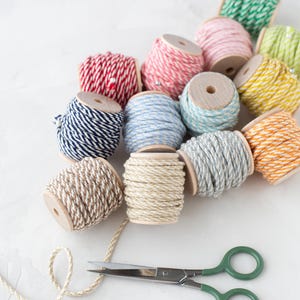 May include: A collection of twelve wooden spools wrapped in multi-colored baker's twine, including red, blue, green, yellow, orange, pink, and neutral tones, arranged with a pair of metal scissors featuring green handles and a single sewing pin.