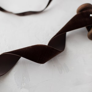 May include: A spool of brown velvet ribbon on a white textured surface. The ribbon is wound around the spool and is partially unwound, creating a soft, flowing curve.