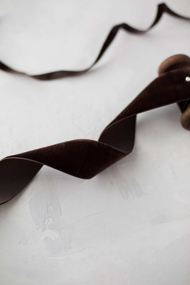 May include: A spool of brown velvet ribbon on a white textured surface. The ribbon is wound around the spool and is partially unwound, creating a soft, flowing curve.
