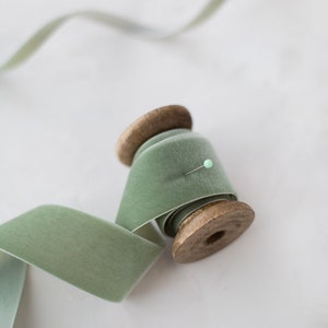 May include: A spool of sage green velvet ribbon with a wooden spool. The ribbon is wrapped around the spool and a pin is holding it in place.
