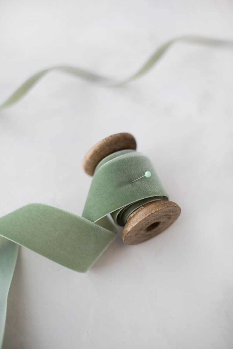May include: A spool of sage green velvet ribbon with a wooden spool. The ribbon is wrapped around the spool and a pin is holding it in place.