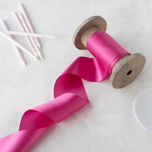 May include: A spool of bright pink satin ribbon on a wooden spool. The ribbon is unraveled and curled on a white surface.