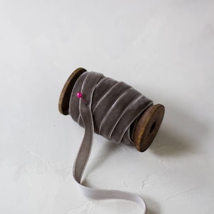 May include: A spool of light gray velvet ribbon with a pink pin on the spool. The ribbon is unfurled and laying on a white surface.