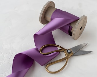 Puede incluir: Un carrete de cinta de satén morado con un carrete de madera y un par de tijeras doradas.