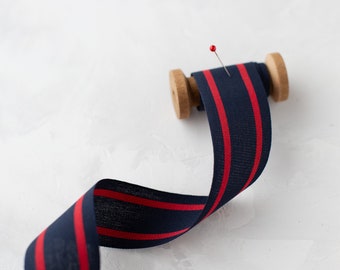 May include: A spool of navy blue ribbon with two red stripes. The ribbon is wound around a wooden spool and a pin is sticking out of the top.