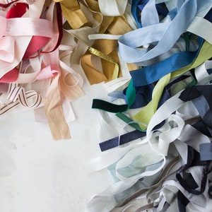 May include: A collection of colorful ribbon scraps in various shades of blue, pink, white, gold, green, and red. The ribbons are arranged in a random pattern on a white surface.