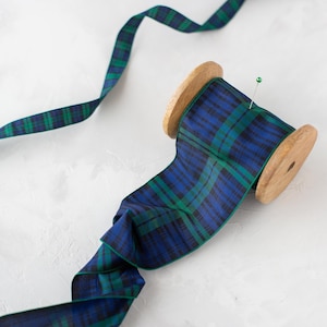 Puede incluir: Un carrete de cinta a cuadros azul y verde con un núcleo de madera. Un alfiler verde asegura la cinta. La cinta está desenrollada, con una parte que se extiende desde el carrete. La cinta mide aproximadamente 5 cm de ancho.