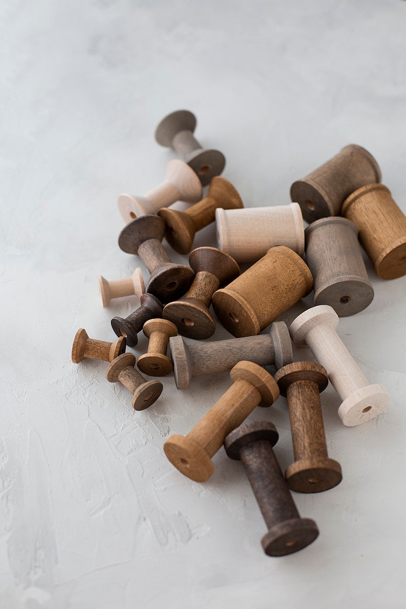 May include: A collection of wooden spools in various shades of brown, beige, and gray. The spools are arranged in a pile on a white surface.