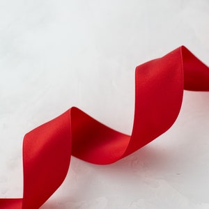 以下が含まれることがあります： 鮮やかな赤いリボンが、質感のある白い表面を優雅にカーブしています。サテンリボンの滑らかな質感が背景と対照的で、視覚的に魅力的なデザインです。ギフト包装やクラフトに最適です。