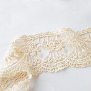 May include: A close-up of a beige crocheted lace trim with a delicate floral pattern. The lace is folded and has a soft, textured appearance.