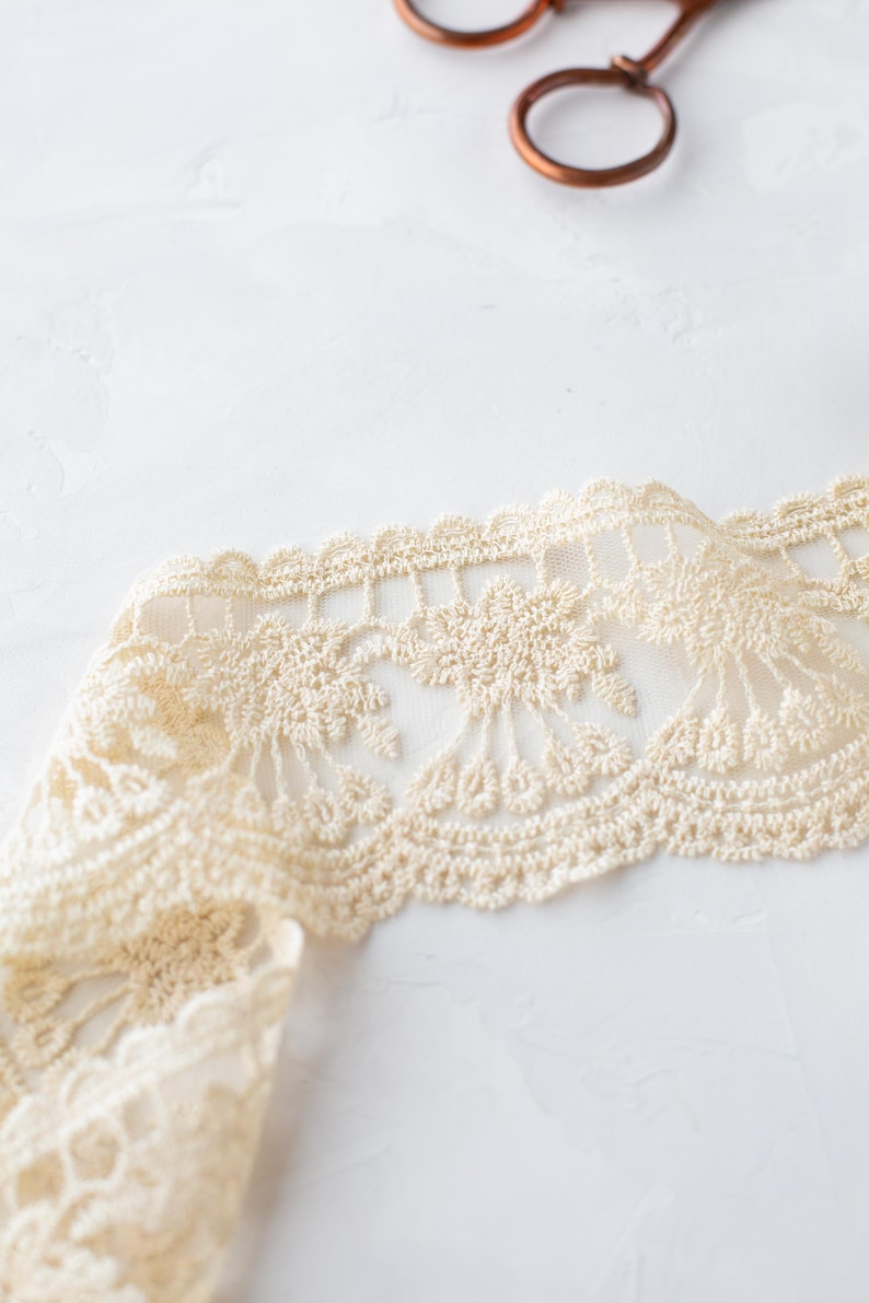 May include: A close-up of a beige crocheted lace trim with a delicate floral pattern. The lace is folded and has a soft, textured appearance.