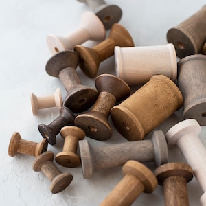 May include: A collection of wooden spools in various shades of brown, gray, and white. The spools are arranged in a pile, with some overlapping. The spools are all different sizes and shapes.