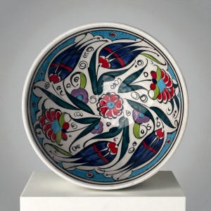 May include: A round ceramic bowl with a white interior and a colorful floral design. The bowl features a blue rim and a central floral motif with red, blue, and green accents. The design includes stylized flowers and leaves.