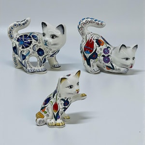 May include: Three ceramic cat figurines. Each cat is white with black facial details and colorful floral patterns in blue, red, and yellow. One cat is standing, one is crouching, and one is sitting with a paw raised.