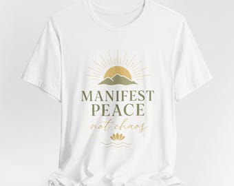 May include: A white short-sleeved t-shirt featuring a graphic design with a gold sun, green mountains, and the text "MANIFEST PEACE not chaos" in green and gold, accompanied by a small lotus flower icon and wavy lines at the bottom.