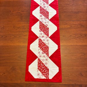 Puede incluir: Camino de mesa acolchado rectangular rojo y blanco. El camino de mesa presenta un diseño central de formas geométricas rojas y blancas alternas. Las secciones blancas están estampadas con motivos florales y de copos de nieve rojos. Los bordes son rojos.