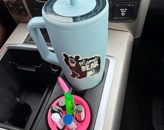 May include: A light blue insulated tumbler with a handle and a straw, featuring a "Mama Bear" sticker. A pink tray holds various small containers and pens. The items are in a car's console.