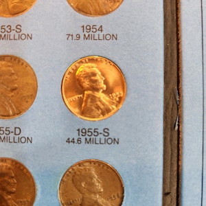 May include: A close-up of a coin collection page featuring several copper-colored Lincoln pennies. The 1955-S penny is prominently displayed, with the text "1955-S 44.6 MILLION" below it. Other coins and dates are also visible.