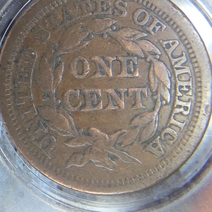 May include: A close-up of a copper-colored United States one-cent coin. The coin features the words "UNITED STATES OF AMERICA" and "ONE CENT" surrounded by a wreath. The coin is encased in a clear protective holder.