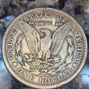 May include: A silver One Dollar coin with the words "UNITED STATES OF AMERICA" and "ONE DOLLAR" encircling an eagle with outstretched wings. The coin also features the phrase "In God we trust".