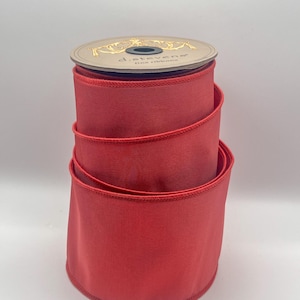May include: A spool of bright red ribbon. The ribbon is tightly wound and has a smooth, satin-like texture. The spool's top is a light tan color with gold lettering and a decorative design. The ribbon is suitable for various crafting and decorative purposes.