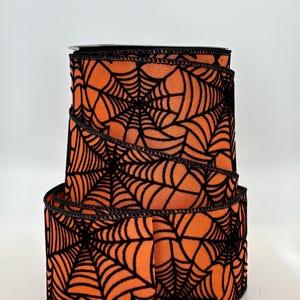 May include: A roll of orange ribbon with a black spiderweb pattern. The ribbon is made of a soft, textured material and is perfect for Halloween or spooky-themed crafts. The edges are finished with a black trim.