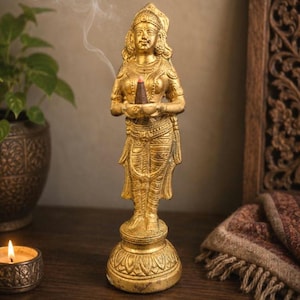 May include: A golden statue of a woman holding an incense cone, with smoke rising. The statue is intricately detailed, standing on a decorative base. A lit candle and a potted plant are in the background, creating a serene atmosphere.