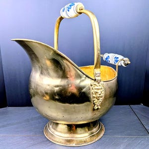 May include: A polished brass coal scuttle featuring a wide, flared opening and a sturdy base. The vessel includes a tall, arched handle and a side handle, both accented with white ceramic grips decorated with blue floral patterns and lion motifs.