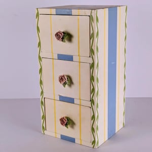 May include: A small, three-drawer cabinet with a decorative floral design. The cabinet features a cream-colored background with yellow and blue vertical stripes. Each drawer has a rose-shaped knob. Green vines with leaves adorn the sides.