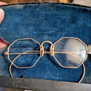 May include: Antique gold-tone eyeglasses with octagonal lenses. The glasses are set in a dark blue velvet-lined case. The frames have a delicate, wire-like design. The glasses are a vintage item.