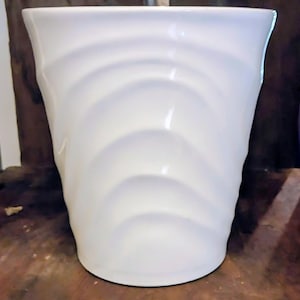 May include: A white ceramic vase with a glossy finish. The vase features a series of curved, wave-like indentations around its exterior. The design gives the vase a textured appearance. The vase is cylindrical and stands upright.