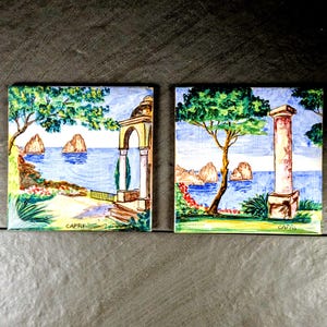 May include: Two square ceramic tiles with hand-painted coastal scenes. One tile depicts a gazebo, the other a column, both overlooking the sea. The tiles feature the word "CAPRI" and are set against a dark background.