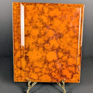 May include: Square orange tray with a marbled pattern and a glossy finish. The tray is supported by a decorative brass stand. The surface has a mottled design in shades of orange and brown, creating a unique visual texture.