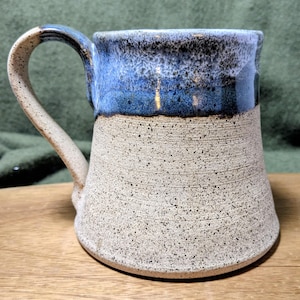 May include: Handmade ceramic mug with a speckled beige base and a blue glazed top. The mug has a large handle and a rustic, textured appearance. Perfect for coffee or tea.