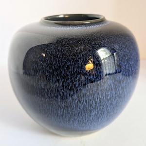 May include: A round, dark blue ceramic vase with a glossy finish. The vase has a speckled texture and a wide opening. The dark blue color fades to a lighter blue near the base. The vase reflects light.