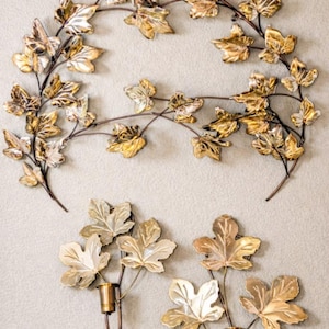 May include: Decorative metal wall art featuring a vine of leaves in gold and silver tones. Below, two candle holders with gold leaves and bronze stems add a touch of elegance. A nature-inspired home decor piece.